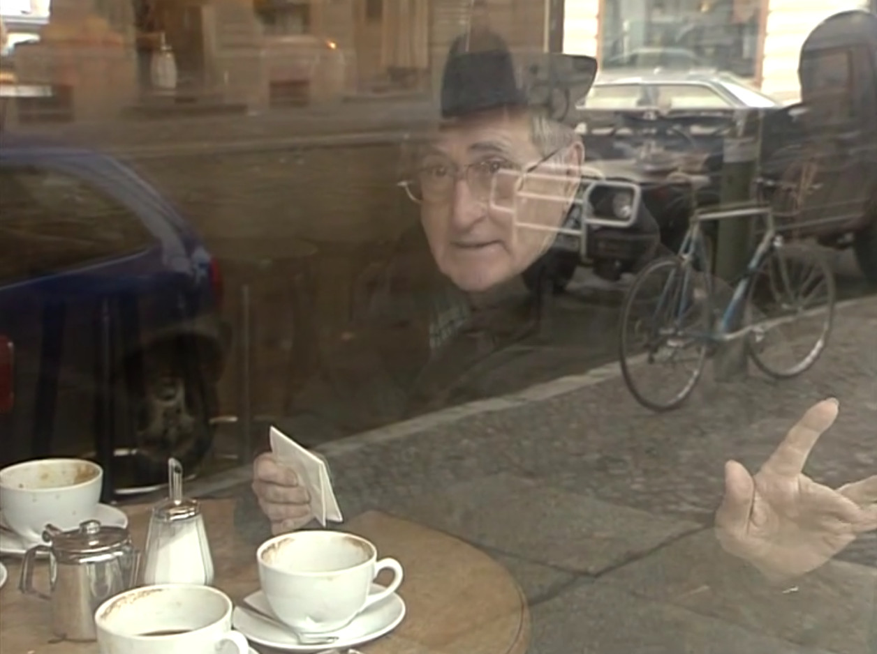 Gad Beck mit Schal und Hut sitzt an einem Bistrotisch mit drei leeren Kaffetassen. Er hält einen zusammengefalteten Zettel in einer Hand, während die andere Hand mit abgespreizten Fingern von sich weist. Gad Beck schaut in die Kamera, die ihn durch das Schaufenster von außen fotografiert.